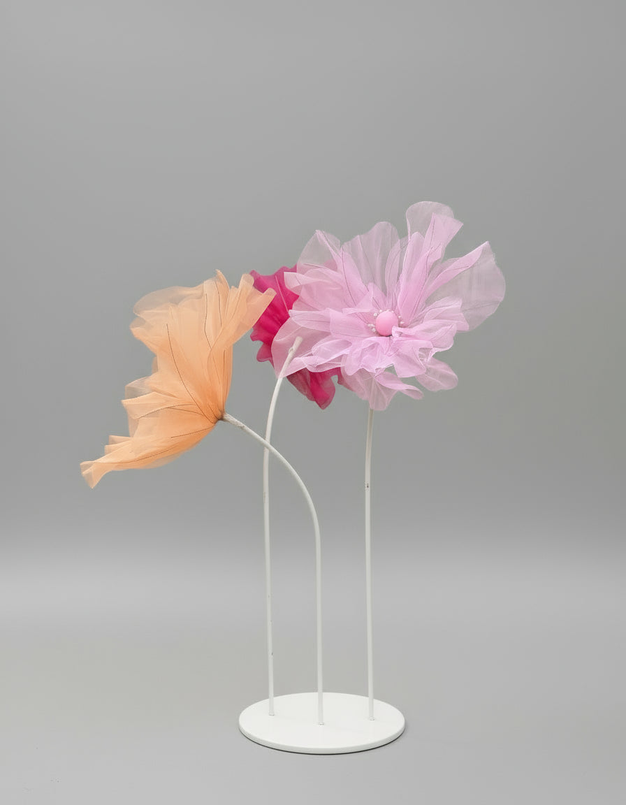 Pink and peach flower-shaped windsocks on stands in a park setting with trees and buildings in the background.